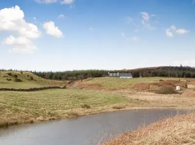 a field with a river and a house on a hill at Cot Cottage - Panoramic Views & Hot Tub! in Castle Douglas