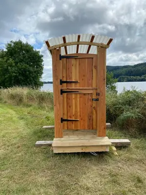 a wooden out house sitting in the grass near the water at The Secret Shepherds Hut- Off Grid Retreat in New Galloway