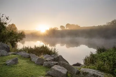 a misty sunrise over a lake with rocks in the foreground at Days Inn Lockerbie - Annandale Water in Johnstonebridge