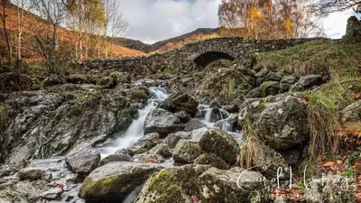 a stream flowing over rocks with a stone bridge at Bakers Cottage in Cockermouth