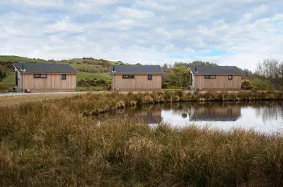 three houses in a field next to a body of water at The Pond Lodges Barstobrick in Ringford